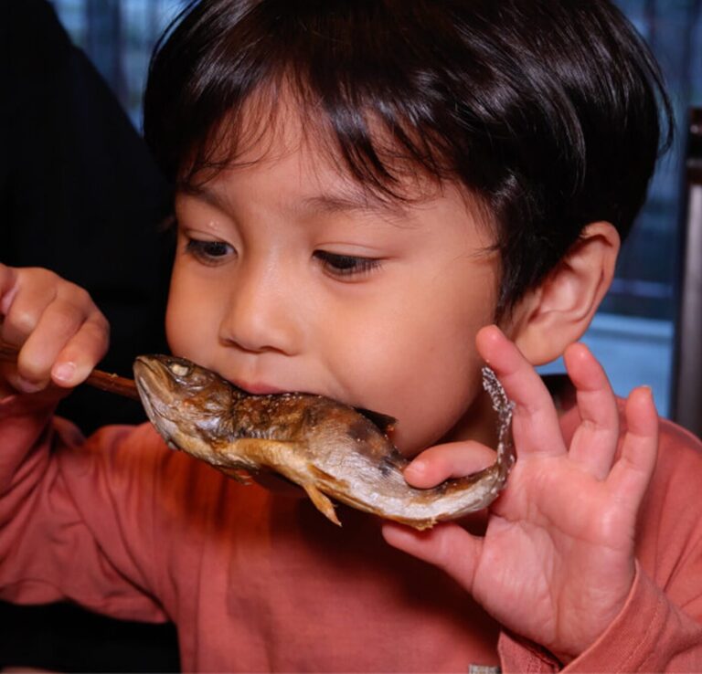 三世代で楽しむ宿のアクティビティで釣った川魚の塩焼きをほおばる男の子。